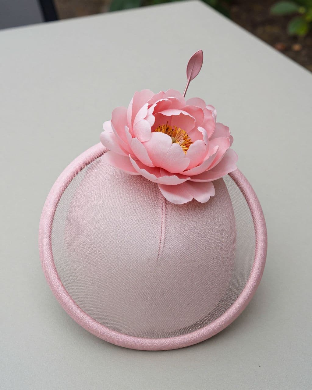 Blush pink fascinator with a looped base and silk peony on a neutral table.