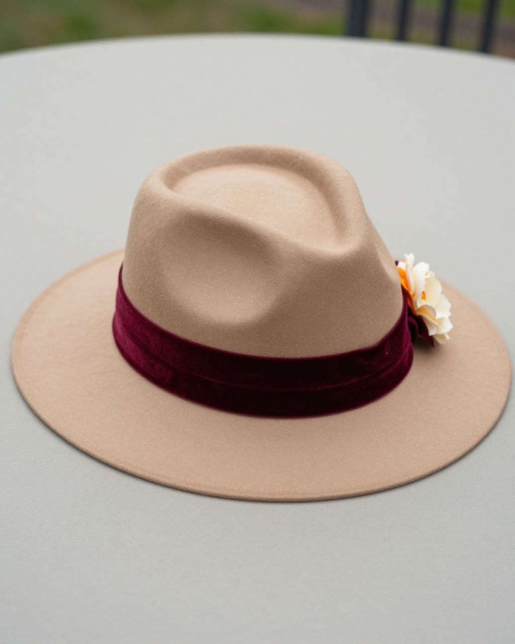 Tan Derby hat with a burgundy velvet ribbon and small cream flower on a tabletop.