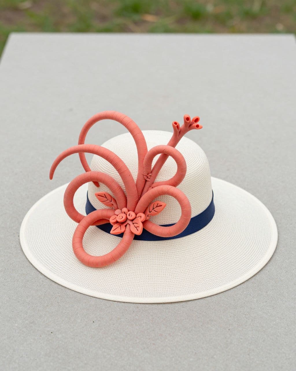 Ivory Derby fascinator with coral loops and navy ribbon on a neutral tabletop.