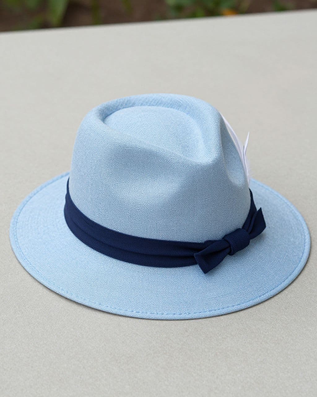 Pale blue sun hat with a linen band, navy knot, and white feather on a tabletop.