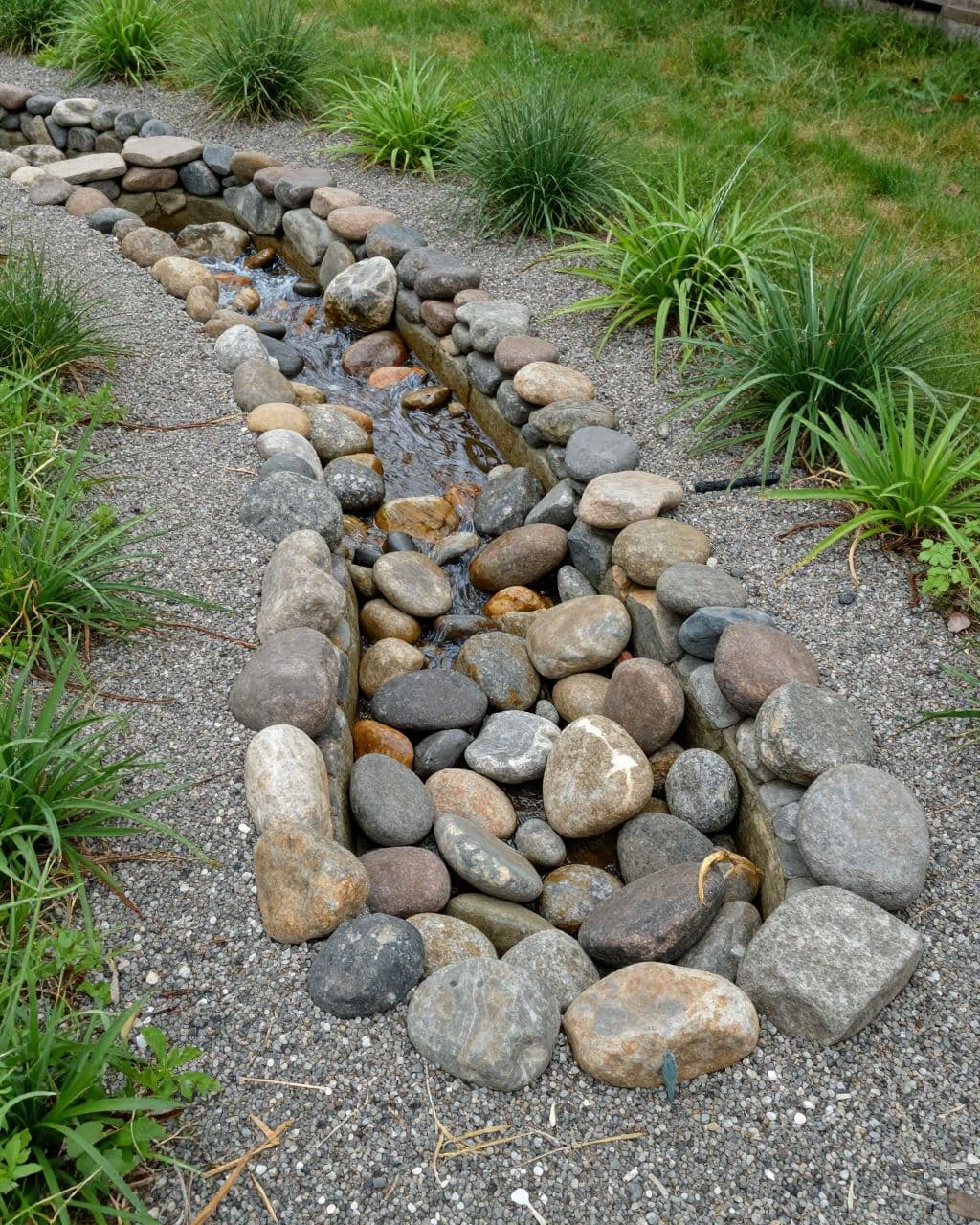 A gravel creek bed leads into a small stone-lined basin beside lawn and native plants.