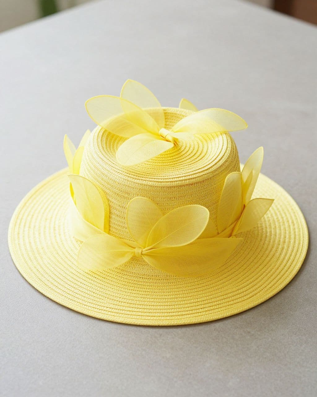 A yellow straw hat with fabric petals around the crown on a neutral tabletop.